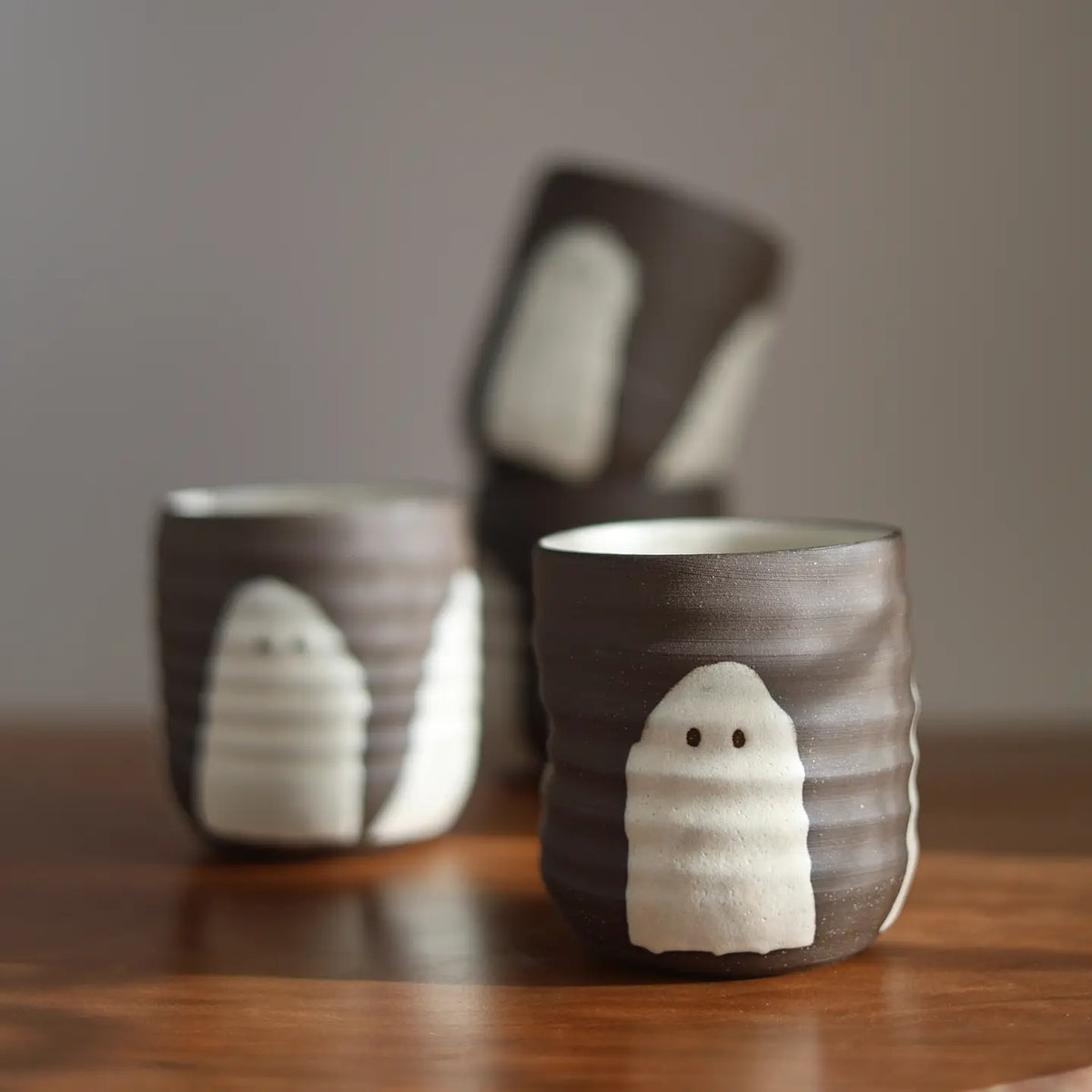 Three dark brown and white ceramic cups with a ghost design, arranged on a wooden table against a neutral gray background.