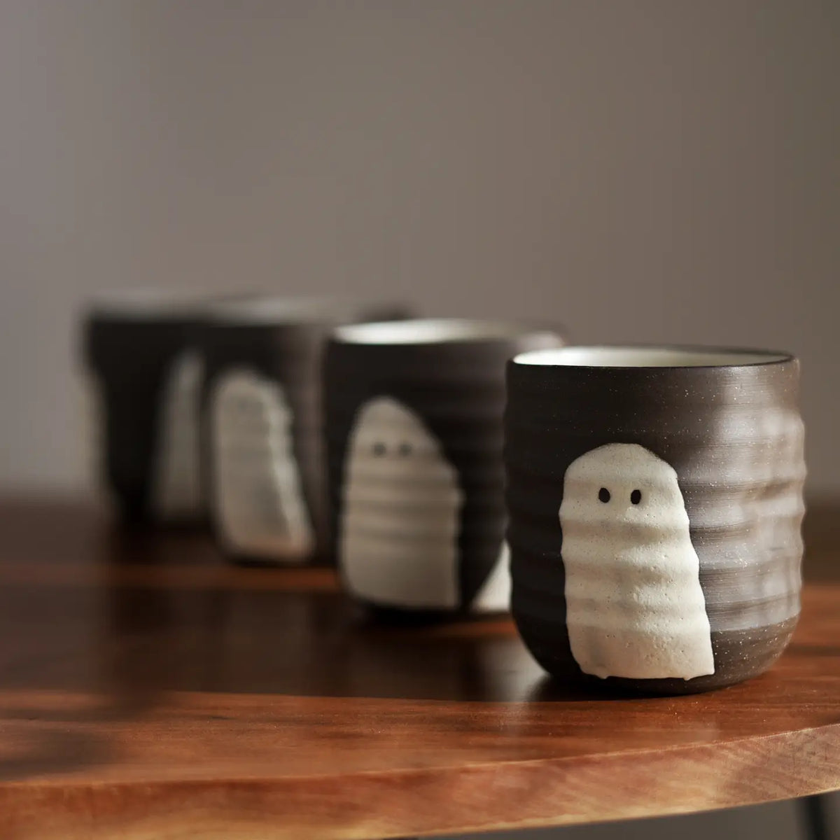 A row of small dark ceramic cups with white ghost-like faces, displayed on a wooden surface against a soft gray background.
