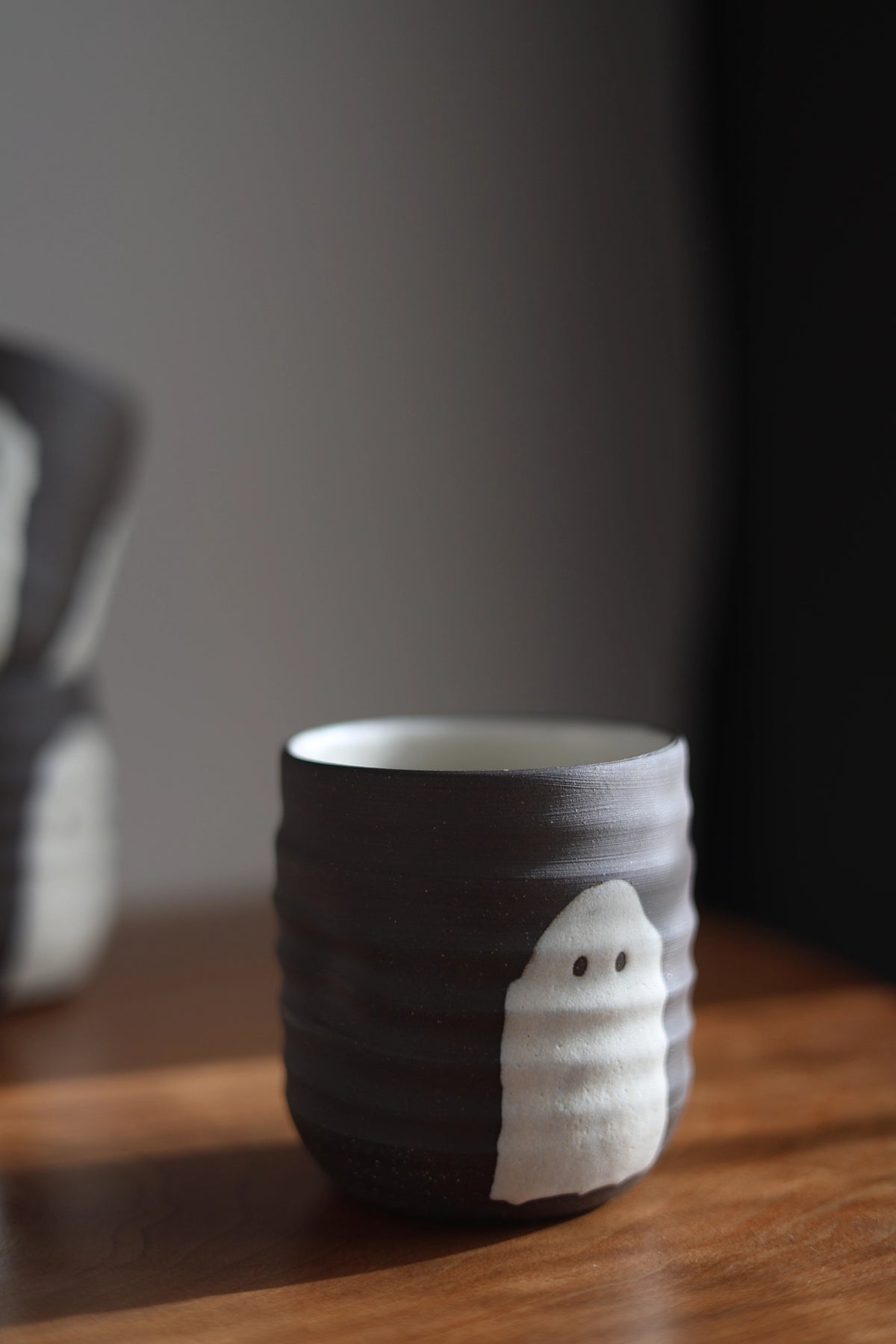 A dark gray ceramic cup with a white ghost-shaped accent and small black eyes, sitting on a wooden surface in a dimly lit setting.