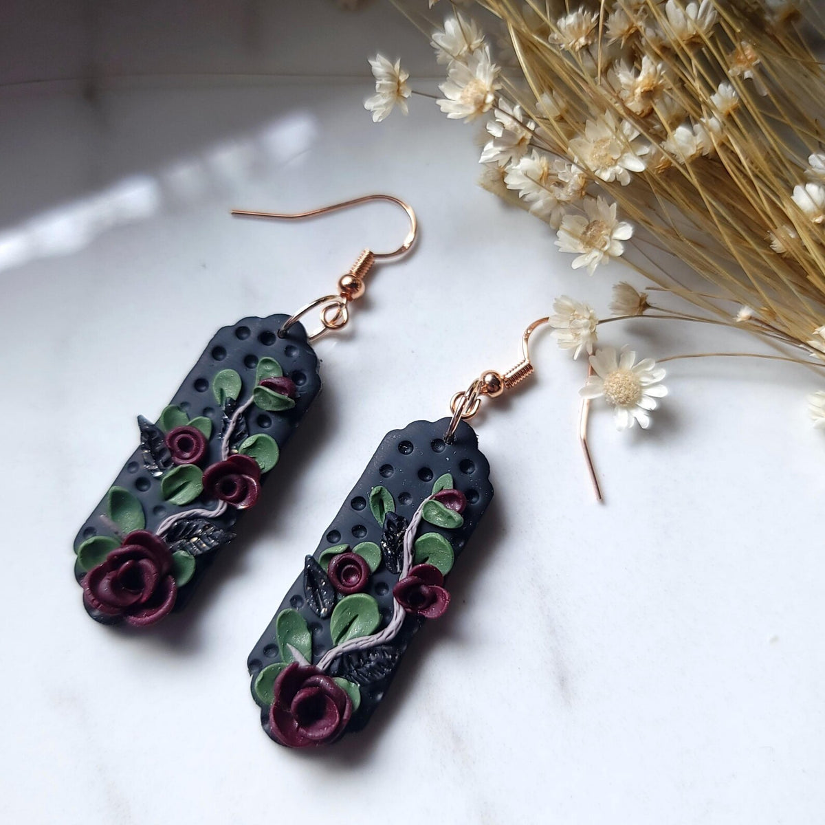 A pair of handmade rectangular drop earrings with a dark charcoal base, featuring raised burgundy roses, green leaves, and silver vine details, hanging from rose gold fishhook wires on a white marble surface with dried flowers.