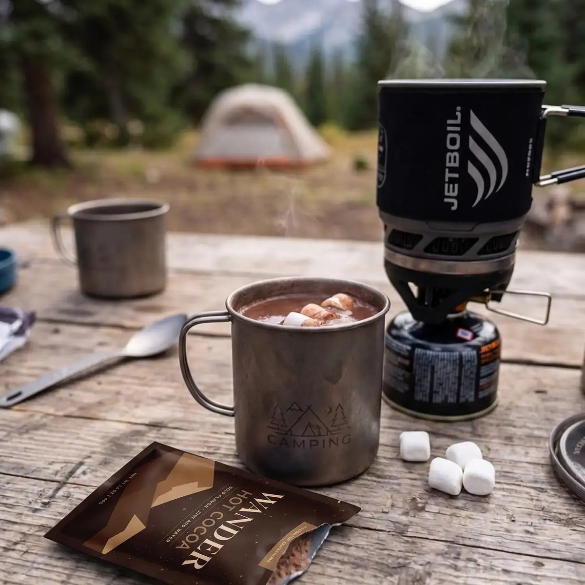 A camping scene on a wooden table with a Jetboil stove, a metal mug of hot cocoa with marshmallows, a packet of Canyon Hot Cocoa, and a blurred campsite background with a tent and trees.