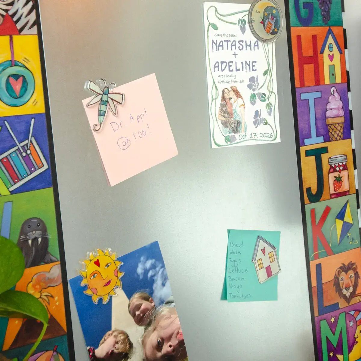 A silver refrigerator door decorated with colorful magnets, a pink sticky note, a printed invitation card, a small teal card, and children's photos, with a child's alphabet poster visible along the right edge.