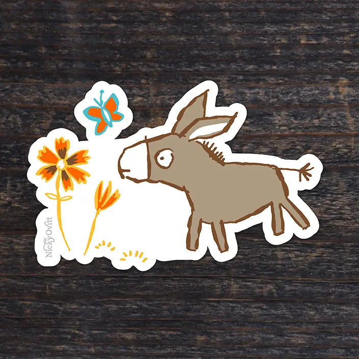 A die-cut sticker featuring a whimsical illustration of a brown donkey, orange flowers, and a blue butterfly on a white background, placed on a dark wood surface. The brand name 'Nico D'Or' is printed vertically on the left edge of the sticker.