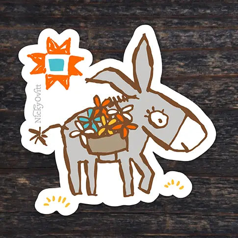 A die-cut sticker featuring a hand-drawn gray donkey carrying a basket of colorful flowers, placed on a dark wood background. The sticker has a white border and includes the brand name 'Nickyovitt' printed vertically on the left side.