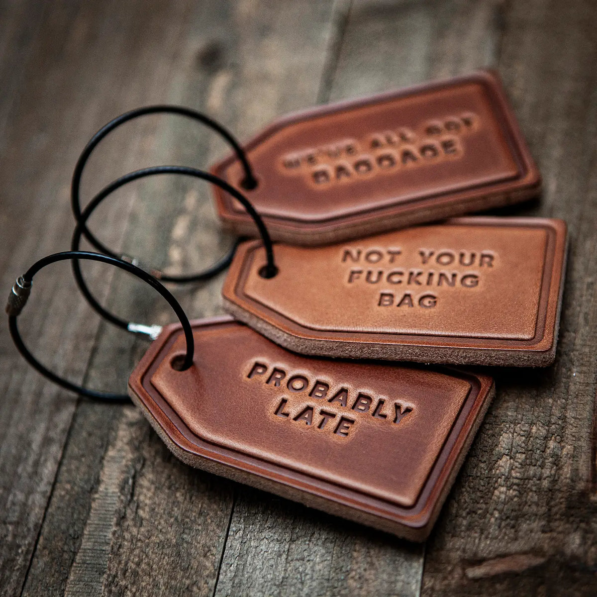 Three brown leather luggage tags with black hanging loops, arranged on a rustic wooden surface. The embossed text reads 'PROBABLY LATE', 'NOT YOUR FUCKING BAG', and 'WE'VE ALL GOT BAGGADE'.