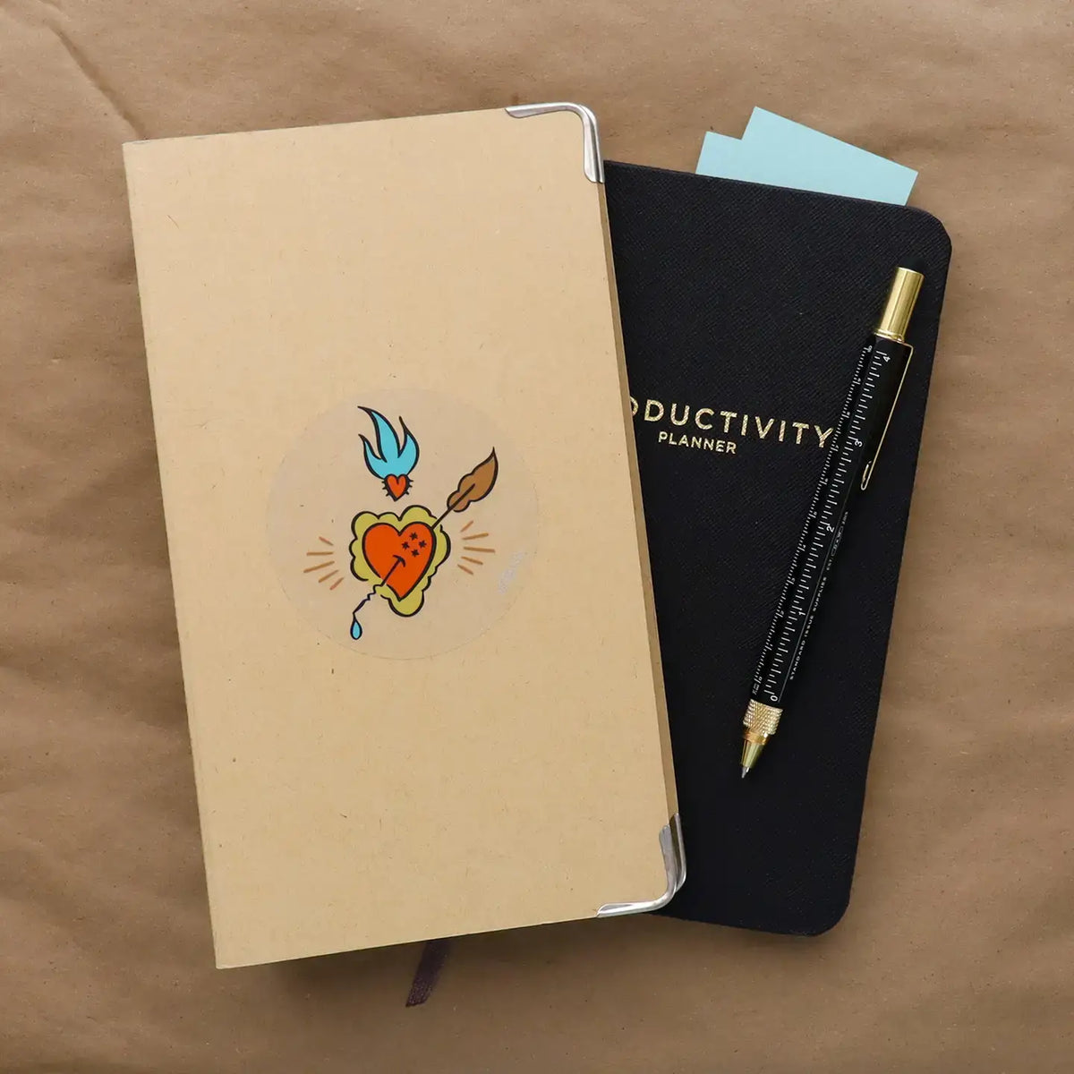 A kraft paper notebook with a heart-and-arrow illustrated sticker sits on top of a black planner labeled 'PRODUCTIVITY PLANNER,' with a black pen featuring gold lettering resting on the planner, all arranged on a brown fabric surface.