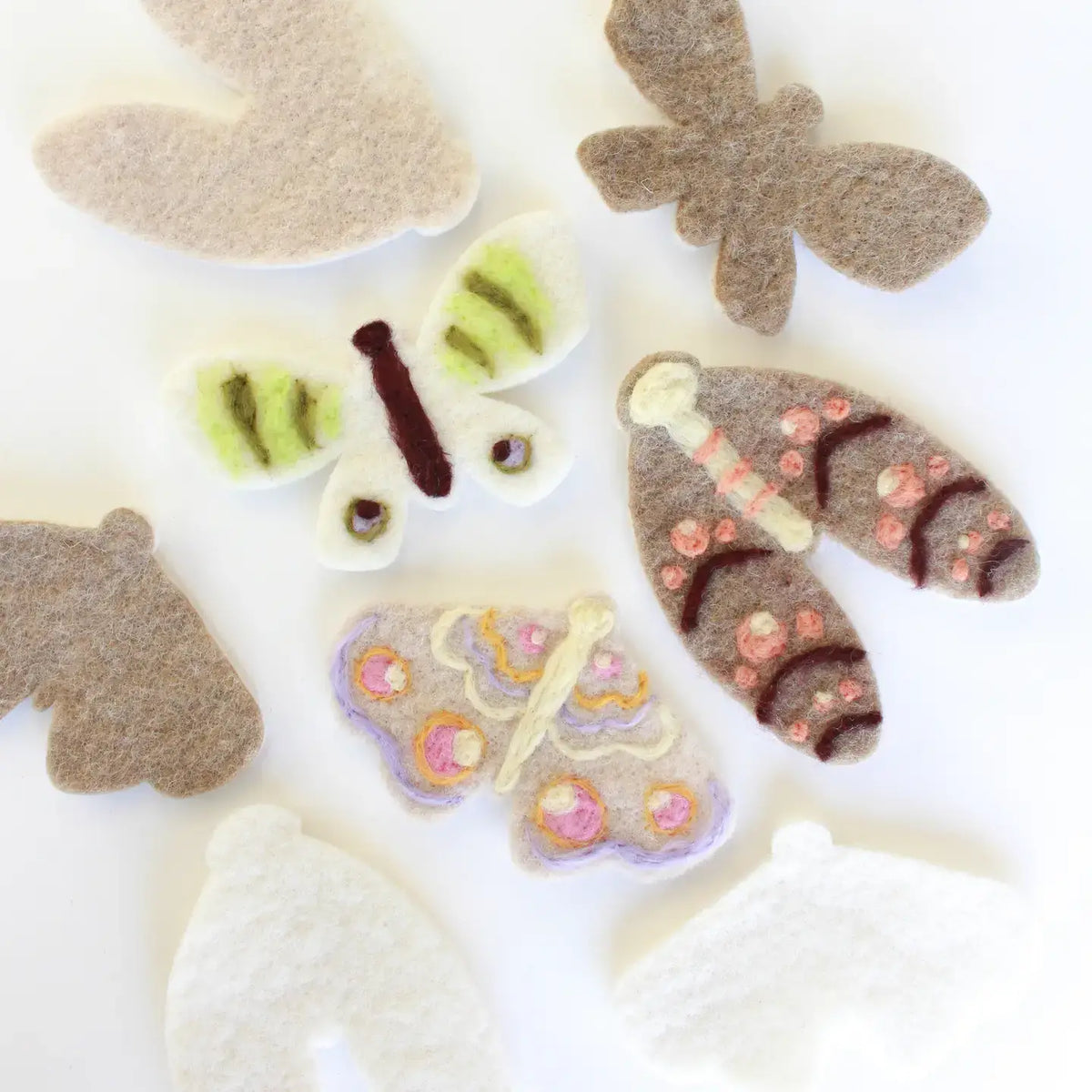 A collection of handcrafted felt moth- and wing-shaped ornaments in various colors, including beige, brown, white, purple, and green, arranged on a plain white background.