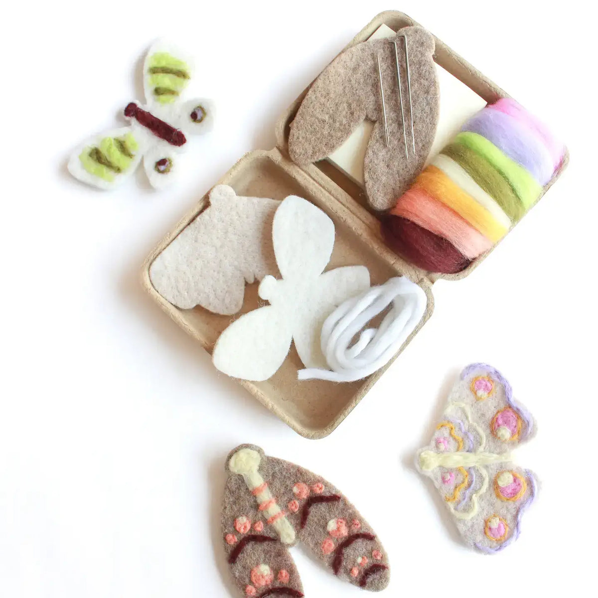A felt moth craft kit arranged on a white background, including felt wings, thread, pins, and a small cardboard tray. Two finished butterfly pieces are shown outside the tray, with one green-and-white butterfly and one lavender-and-pink butterfly with floral details.
