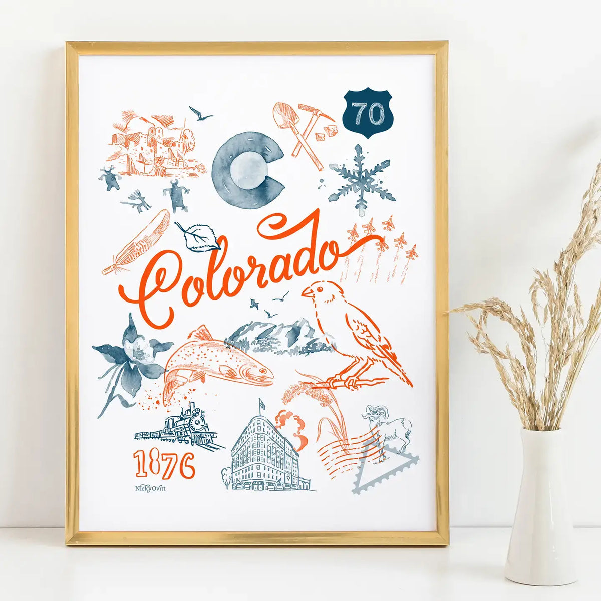 A framed Colorado-themed art print with orange and blue illustrations, including the word 'Colorado' in script, a Route 60 shield, mountains, a bird, and '1876' text, displayed on a white surface with a vase of dried grass beside it.