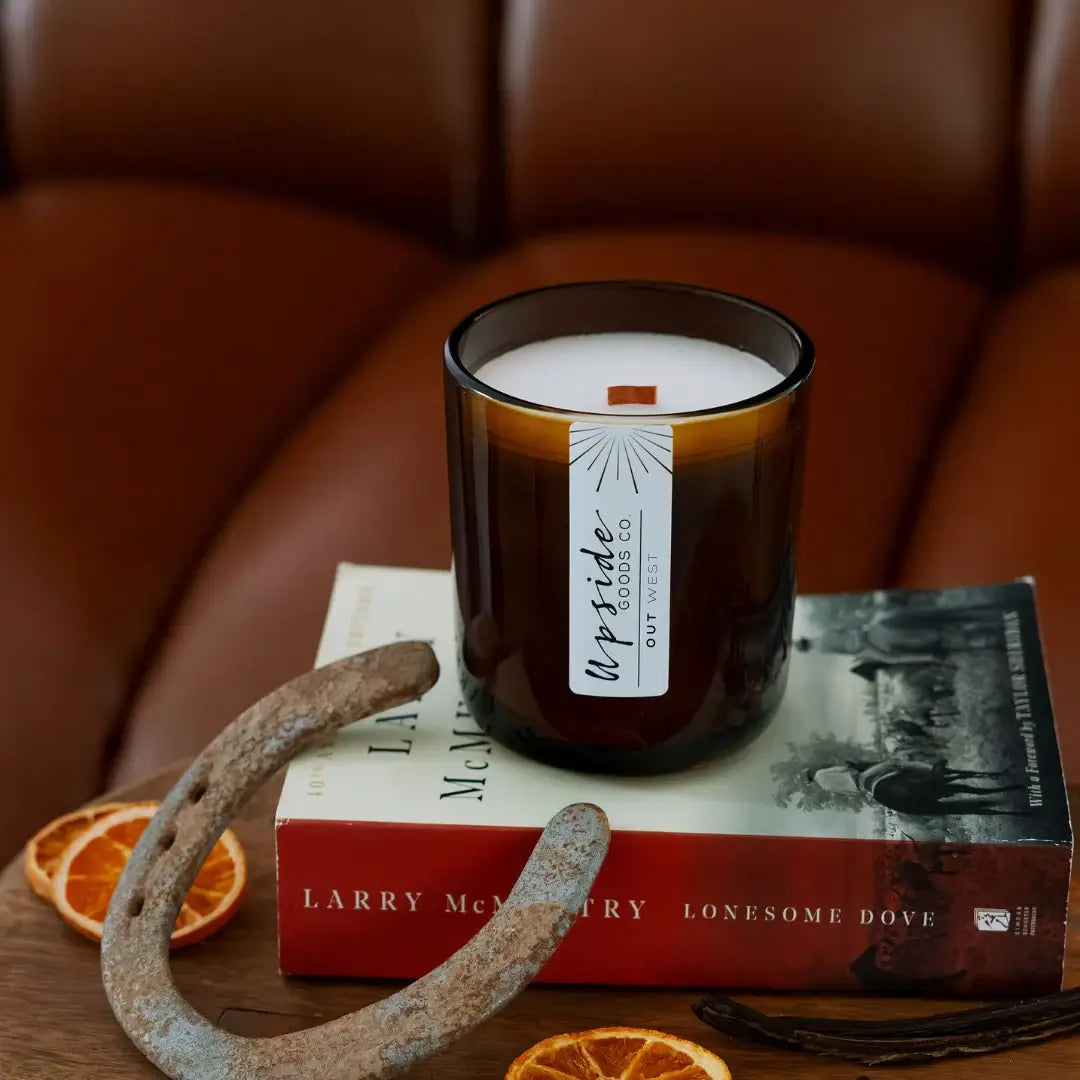 A candle in a dark amber glass jar sits on top of a book, styled with dried orange slices, a rusted horseshoe, and vanilla beans in front of a brown leather chair. The candle label reads 'Upside Goods Out West'.