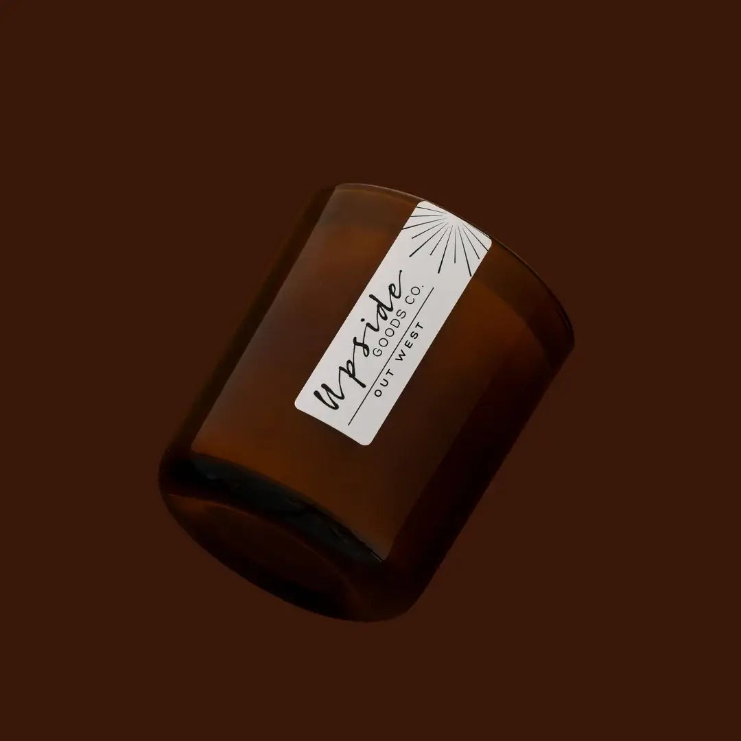 An amber glass jar candle with a white paper label reading 'Upside Candle Co. Out West' on a solid dark brown background.