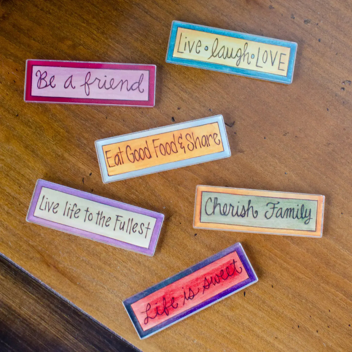 Six rectangular decorative magnets with colorful borders and inspirational hand-lettered quotes, arranged on a wooden tabletop.
