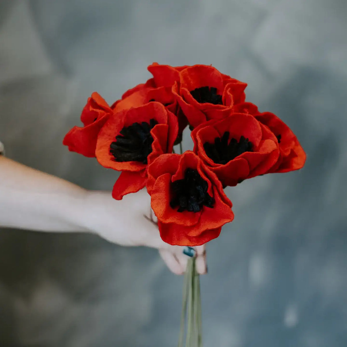 bouquet of red felted poppies, green floral wire stems being held by a hand with grey background
