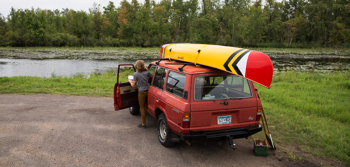 Red van with a yellow Sanborn Canoe Co. kayak on top, parked by a lake with a person getting inside.