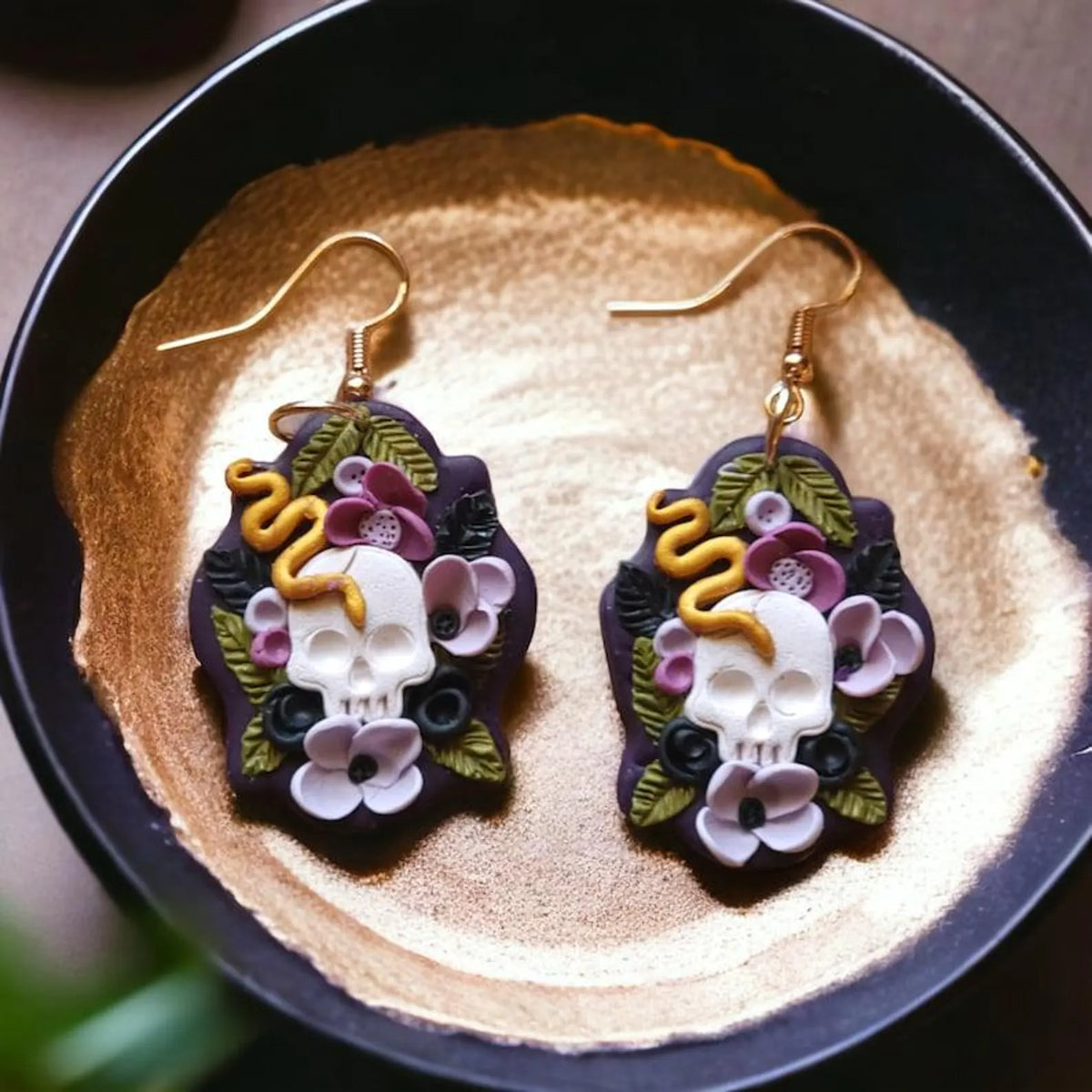 A pair of gothic dangle earrings featuring white skulls, pink flowers, green leaves, and a yellow snake on a dark purple background, displayed on a textured gold surface inside a black bowl.