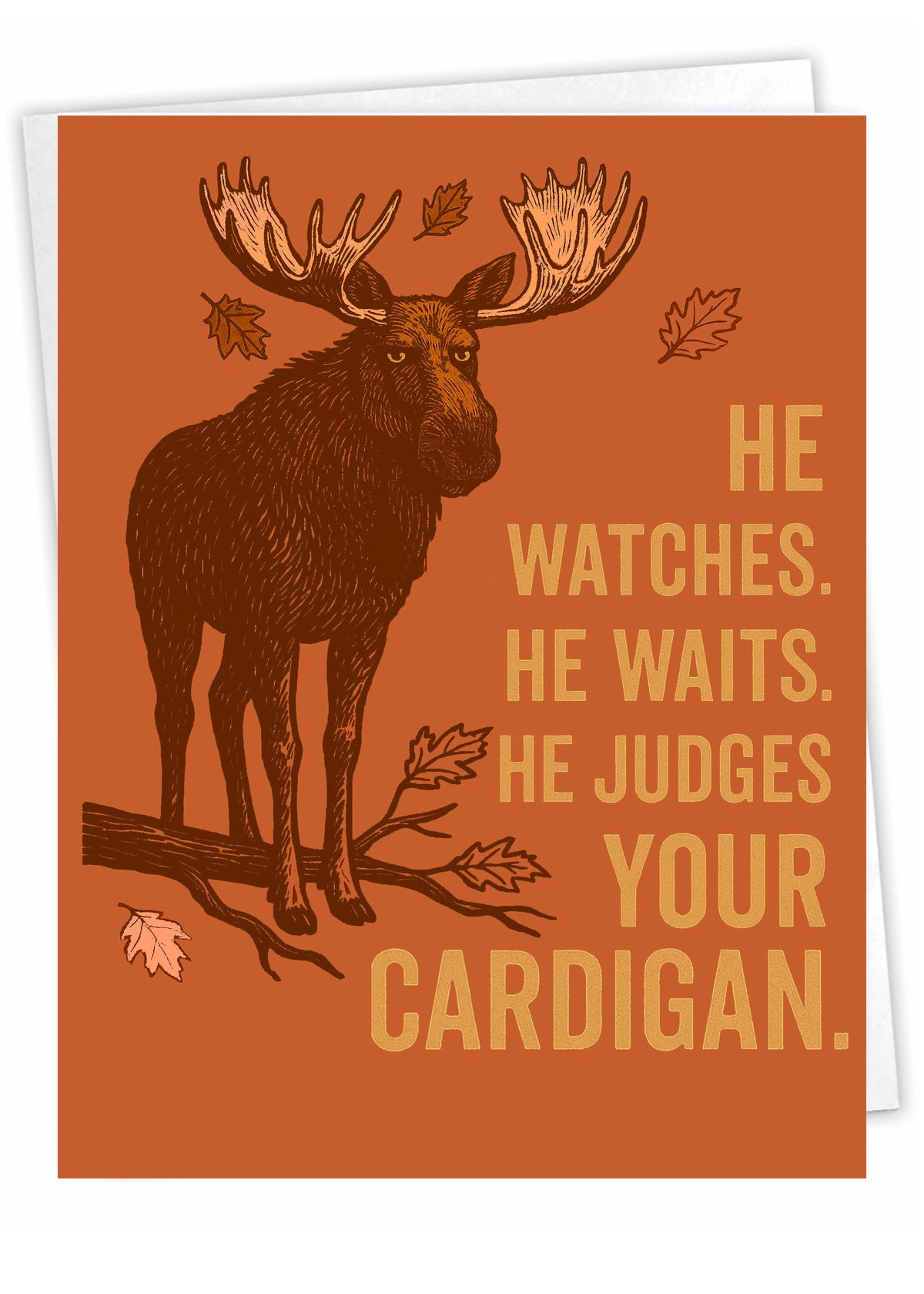 A rectangular greeting card or print with an orange-brown background, featuring a dark brown moose illustration with large antlers standing on a branch, plus scattered autumn leaves and the text: 'HE WATCHES. HE WAITS.