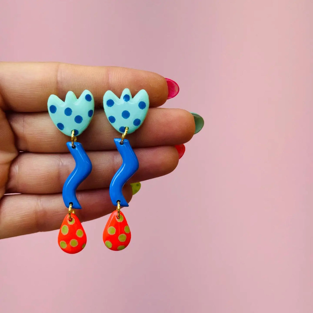 A hand with colorful painted fingernails holds a pair of handmade dangle earrings against a soft pink background. The earrings feature light blue tulip-shaped tops with dark blue polka dots, connected to bright blue wavy stems, and ending in red teardrop charms with yellow polka dots.
