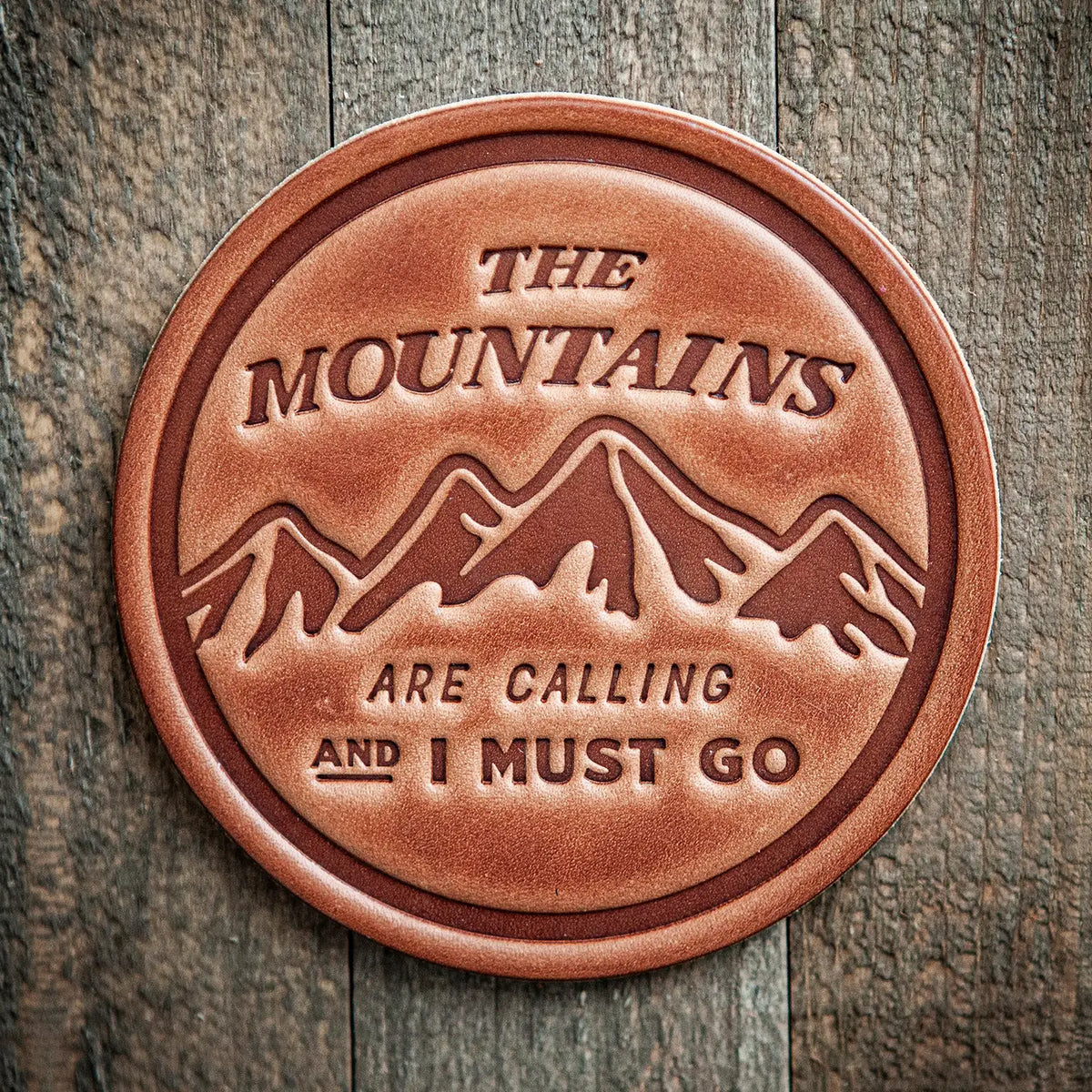 A round leather coaster or plaque with an embossed mountain design and the text 'THE MOUNTAINS ARE CALLING AND I MUST GO,' shown on a rustic wood plank background.