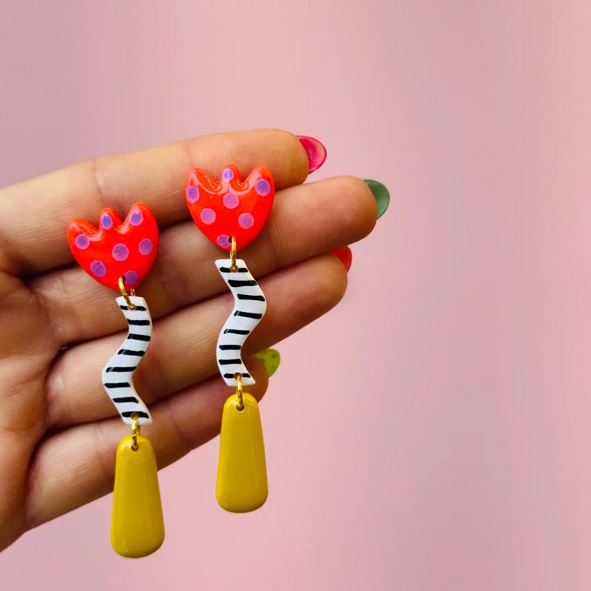 A close-up of a hand holding a pair of colorful dangling earrings against a soft pink background. The earrings feature red heart-shaped studs with purple dots, black-and-white wavy strips, and yellow teardrop pendants.