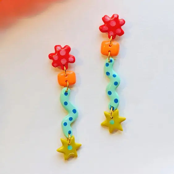 A pair of colorful dangling earrings with red flower charms, orange beads, a mint green wavy bead with blue dots, and yellow star charms, displayed on a white surface.