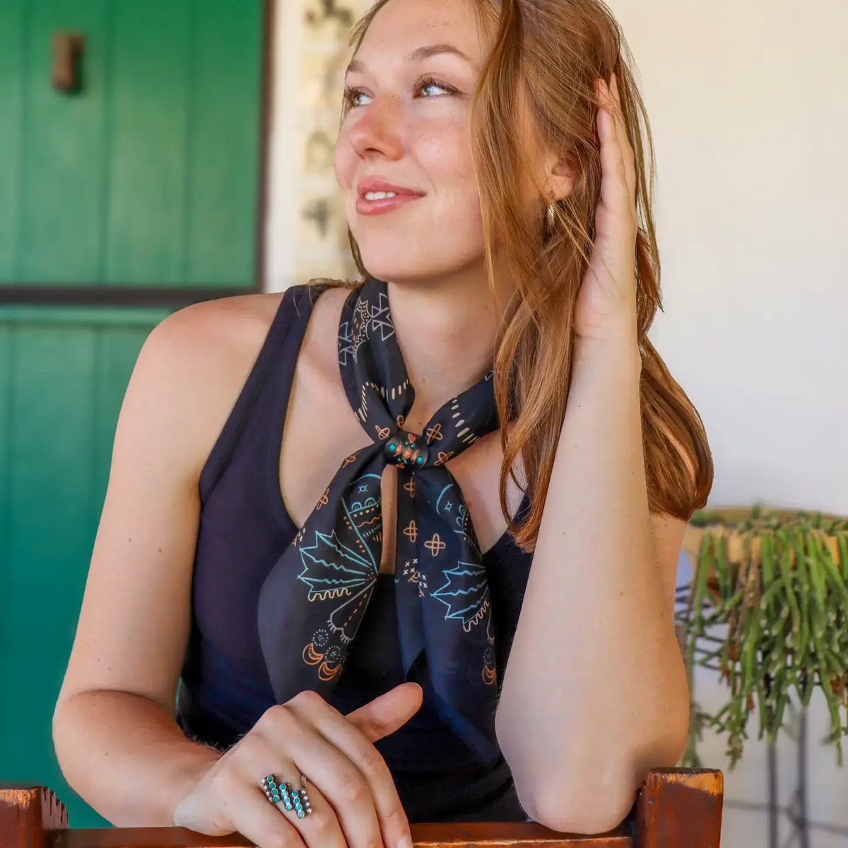 A woman wearing a navy blue silk scarf with a bohemian pattern, tied around her neck over a dark top, in a brightly lit indoor setting.