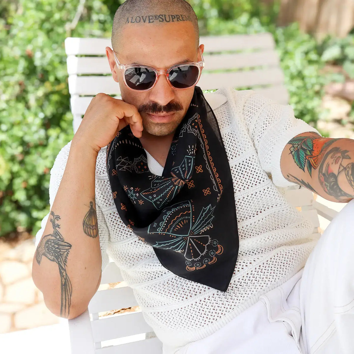 A man wearing a blue bandana-style neck scarf with colorful butterfly and floral patterns, styled with sunglasses and a white knit shirt while sitting on a white outdoor chair.