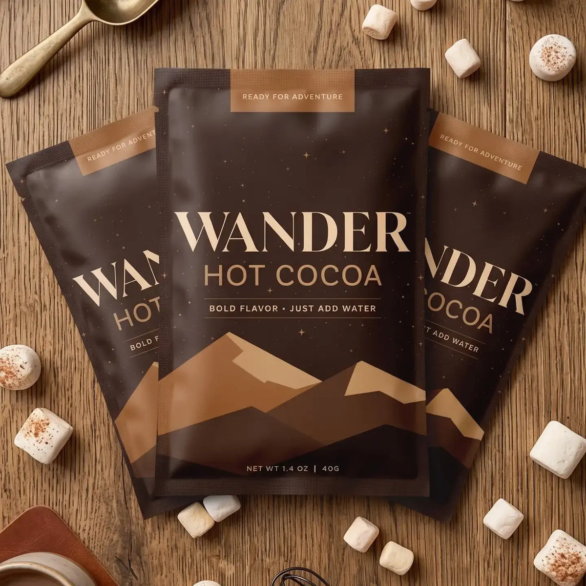 Three bags of Wander Hot Cocoa arranged on a wooden surface with marshmallows and a spoon around them. The center bag is fully visible, with two matching bags partially behind it on both sides.