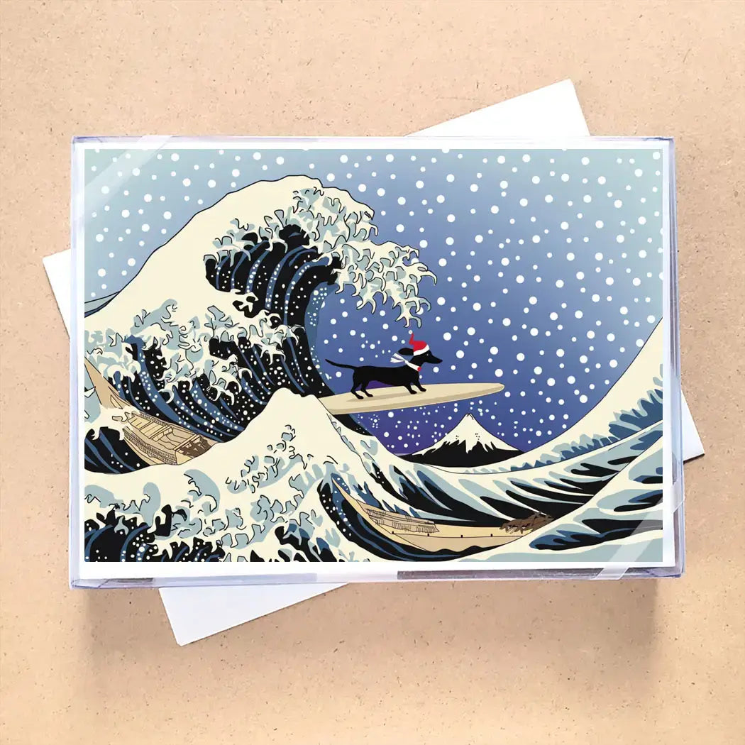 A square greeting card or art print featuring a stylized blue and white wave scene with snow, a small black dog on a surfboard, and a Santa hat, photographed on a beige surface with white envelopes behind it.