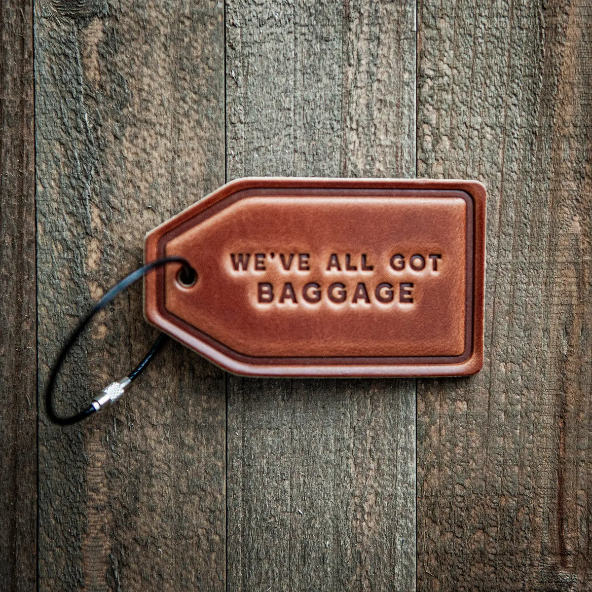 A brown leather luggage tag with the embossed text 'WE'VE ALL GOT BAGGAGE' and a black loop, shown on a rustic dark wood background.