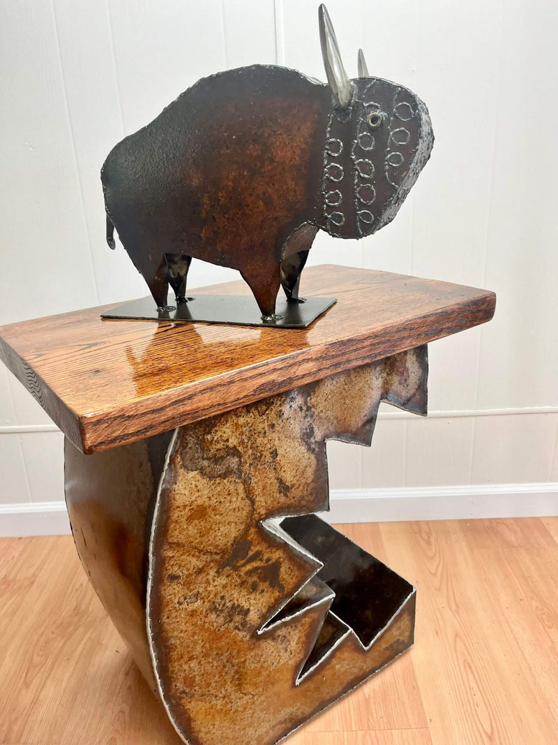A rustic metal buffalo sculpture displayed on a wooden and metal side table against a white paneled wall and light wood floor.
