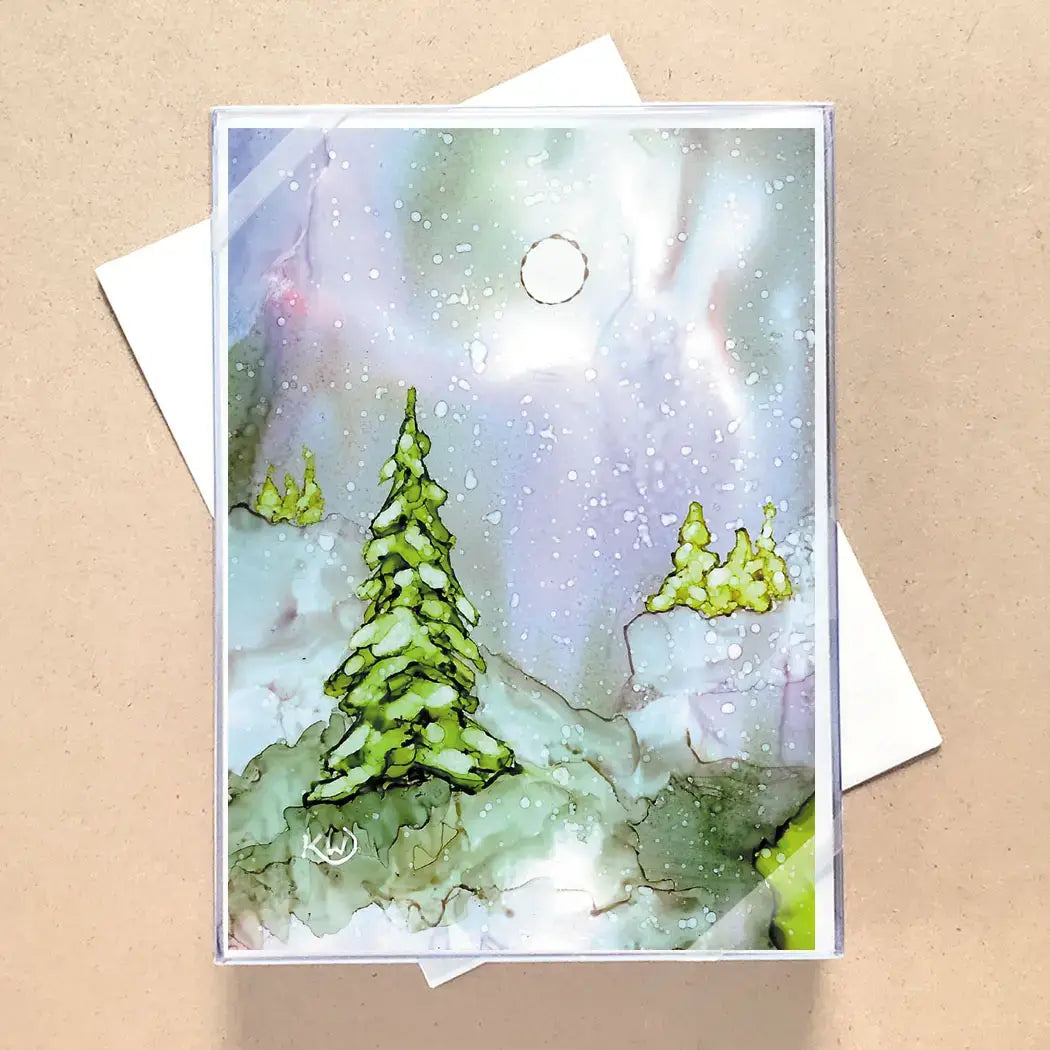 A square greeting card with a watercolor winter landscape design, featuring green pine trees under a soft pastel sky, displayed on a kraft paper background with a white envelope behind it.