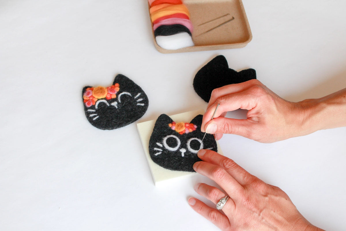 Black cat-shaped felt decorations with floral details on a white surface, with a hand interacting with them.