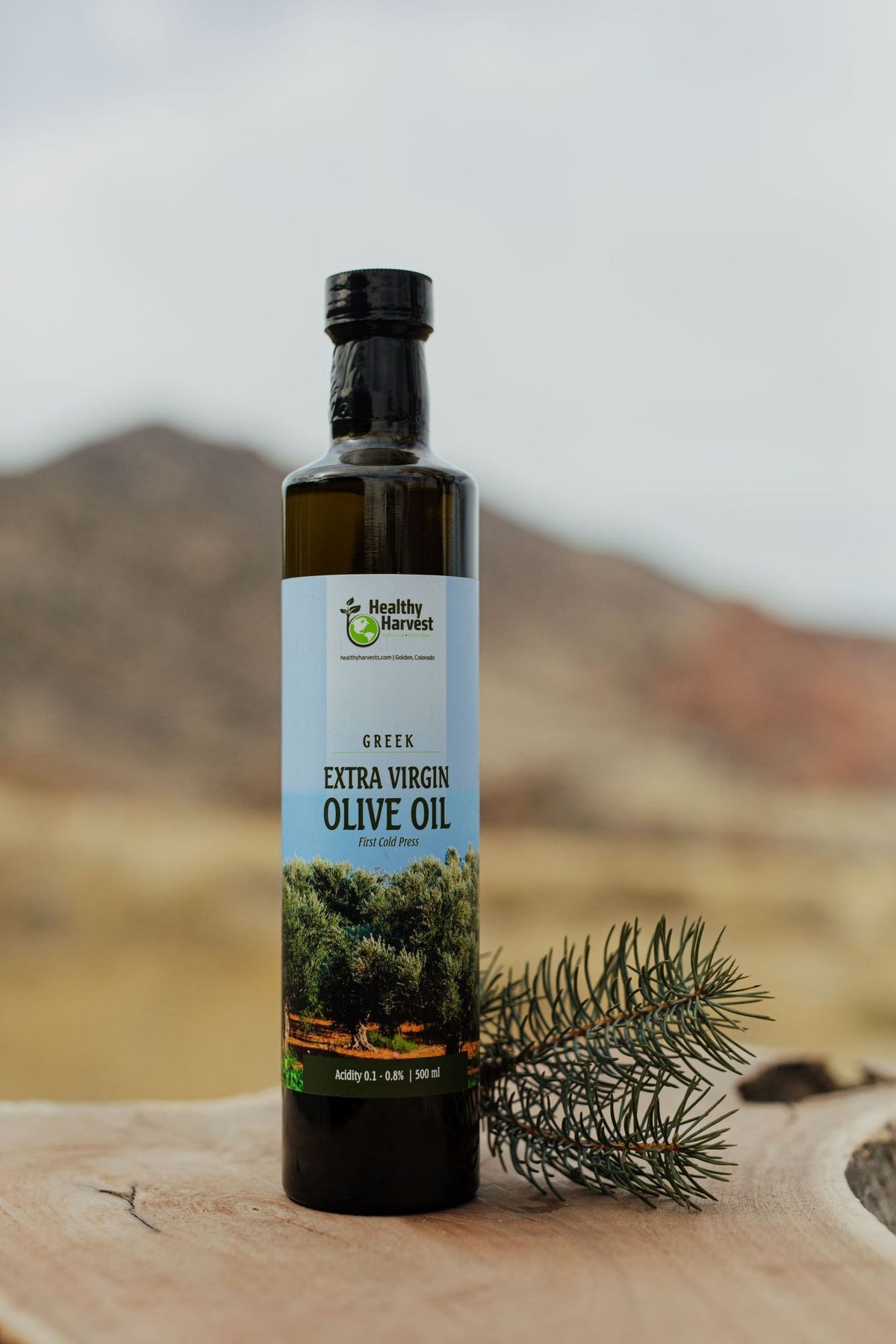 500mL Bottle of Healthy Harvest Extra Virgin Olive Oil photographed outside with a  branch and mountain in background highlighting the bright blue sky and Greek olive farm on the label