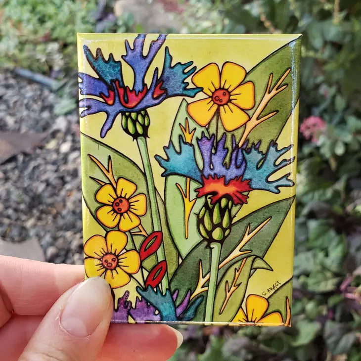A hand holding a colorful refrigerator magnet with a floral cornflower and mountain bluet design, featuring flowers in yellow, blue, and red colors.