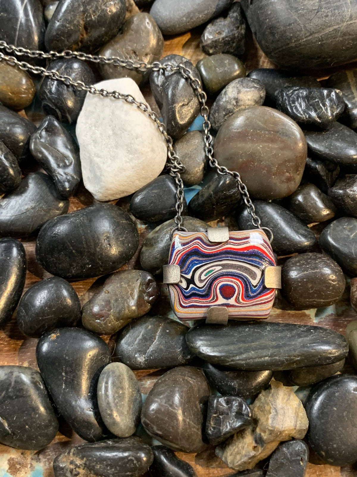 black river rock background with a one of a kind square red, white and blue fordite cabachon set with oxidized sterling silver tabs on a 16" chain
