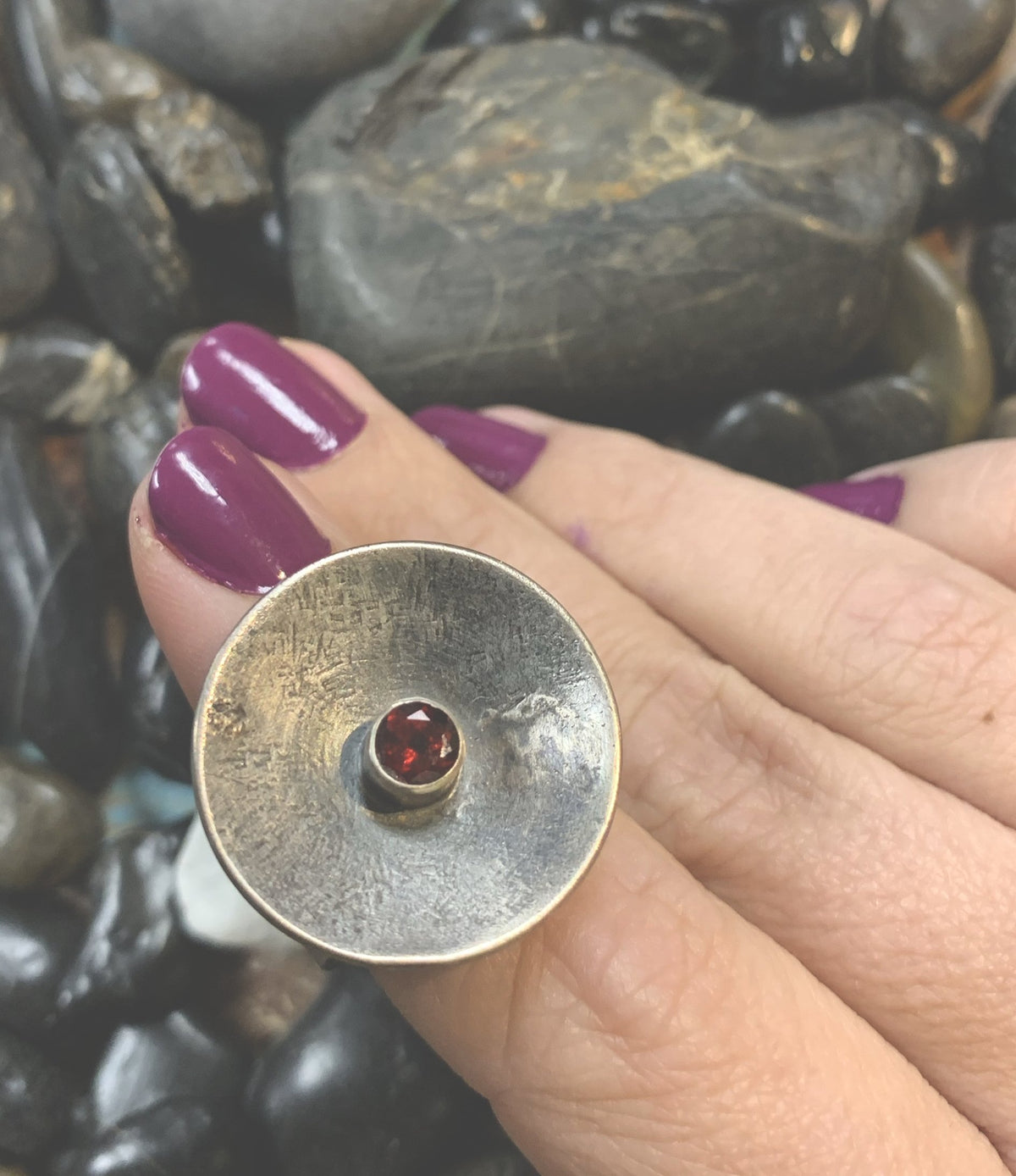 Sterling Silver disk ring with a small garnet set in the center shot on a white womans hand woth purple nail polish